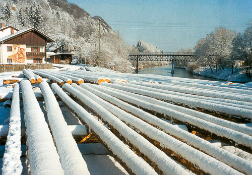 Verschneite Baumstämme für den Flossbau lagern im Winter an der Loisach