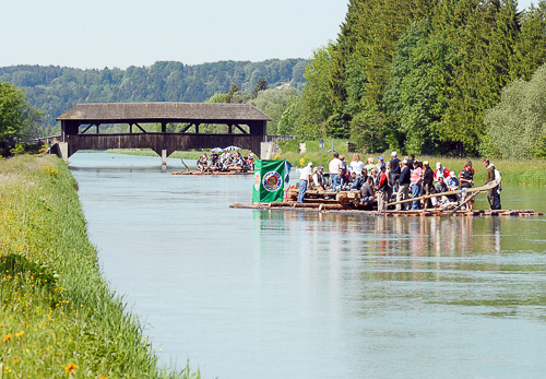 Zwei Flöße passieren eine historische Holzbrücke auf der Isar bei Sonnenschein